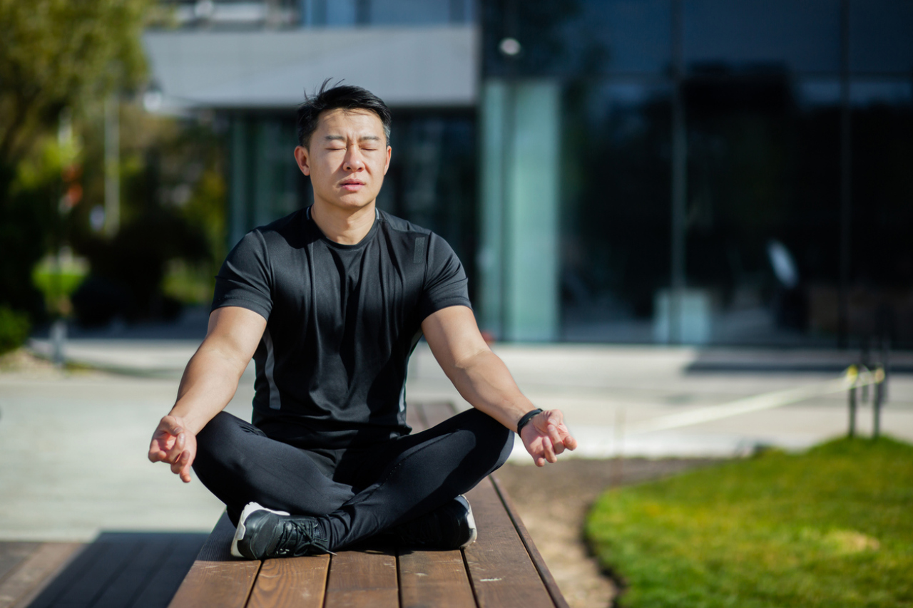 一位穿著黑色運動服的男子坐在戶外的木製長椅上，雙腿盤坐，雙手放在膝蓋上，眼睛閉著在冥想