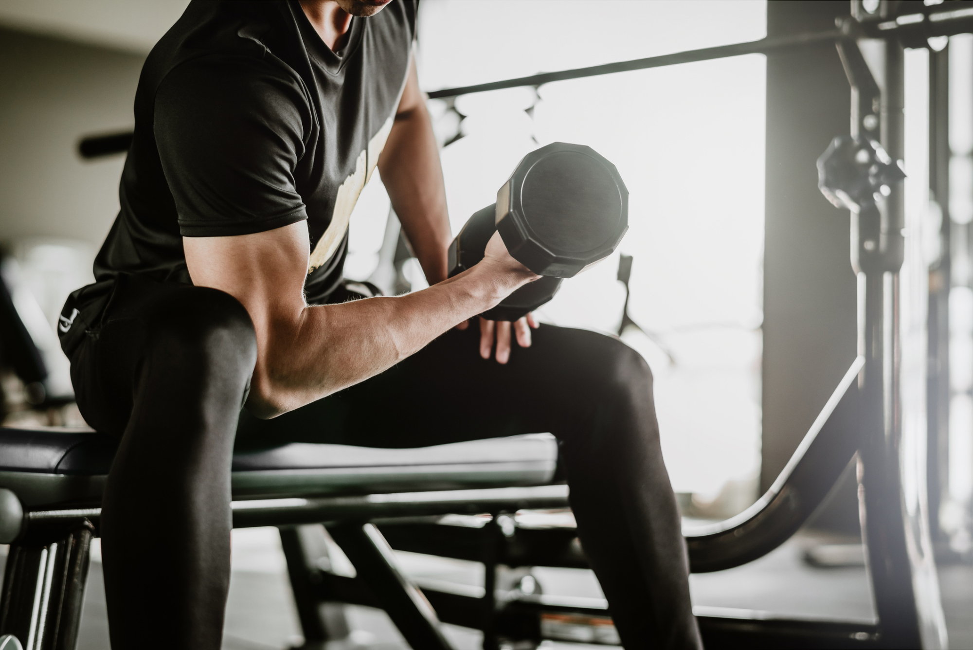 A close-up of a man sitting on a weight bench in a gym, performing a bicep curl with a dumbbell.