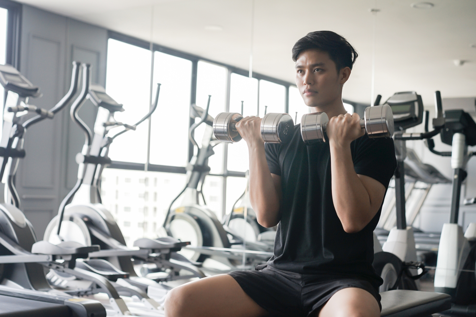  A young man sitting on a machine in a gym, holding a dumbbell in each hand and performing bicep curls.