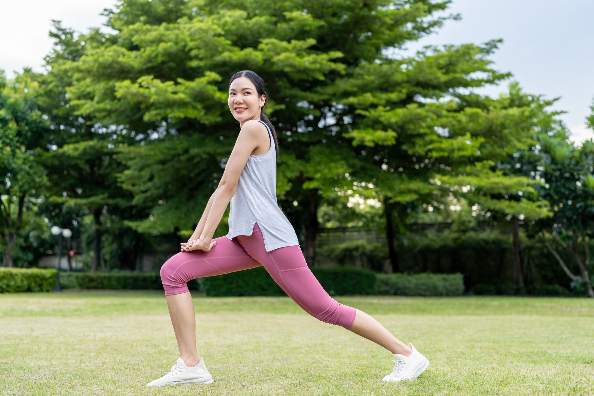 iStock-2178368968.jpg A woman doing lunge stretches on park grass, stepping forward and squatting to stretch hip flexors and front thigh muscles.