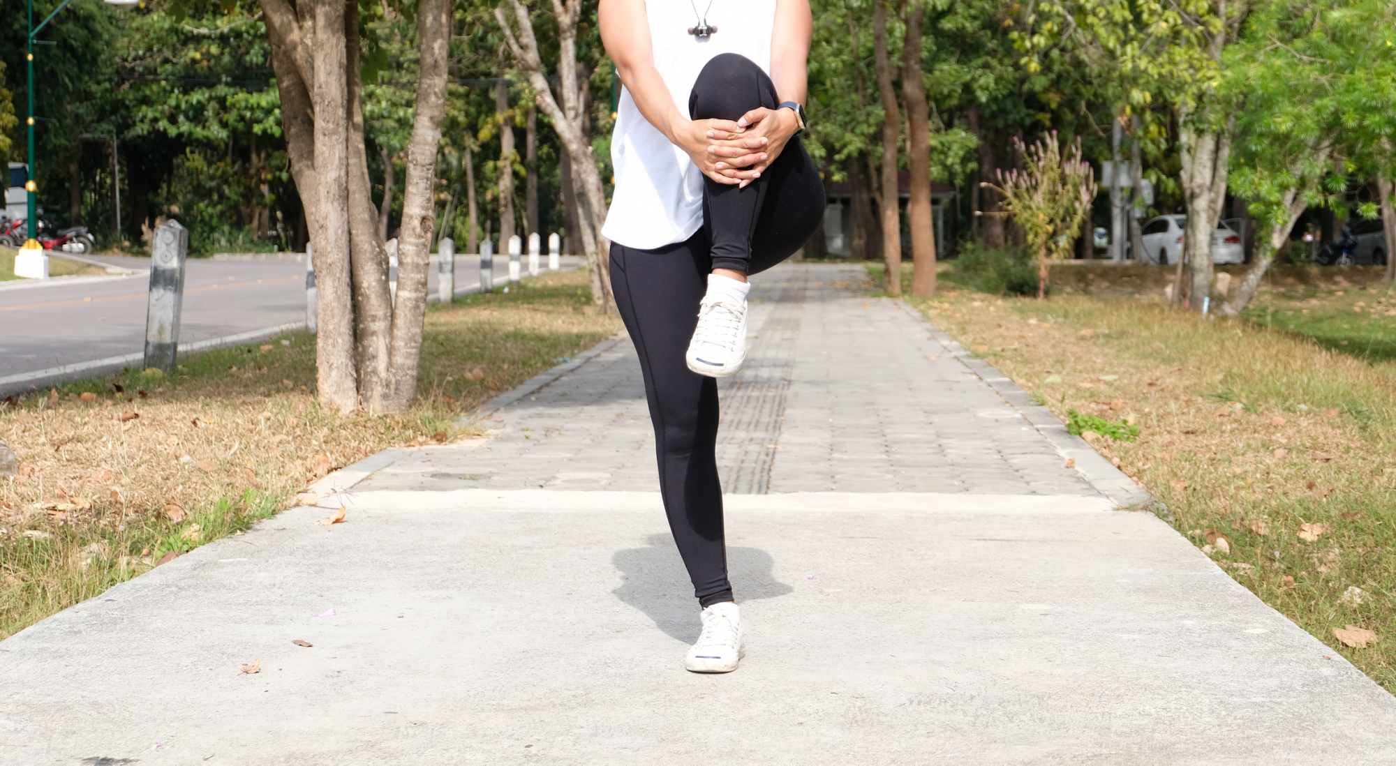 iStock-1580791641.jpg A woman doing knee hug dynamic warm-up outdoors, standing on one leg and hugging the knee to the chest to stretch glutes and hip joints
